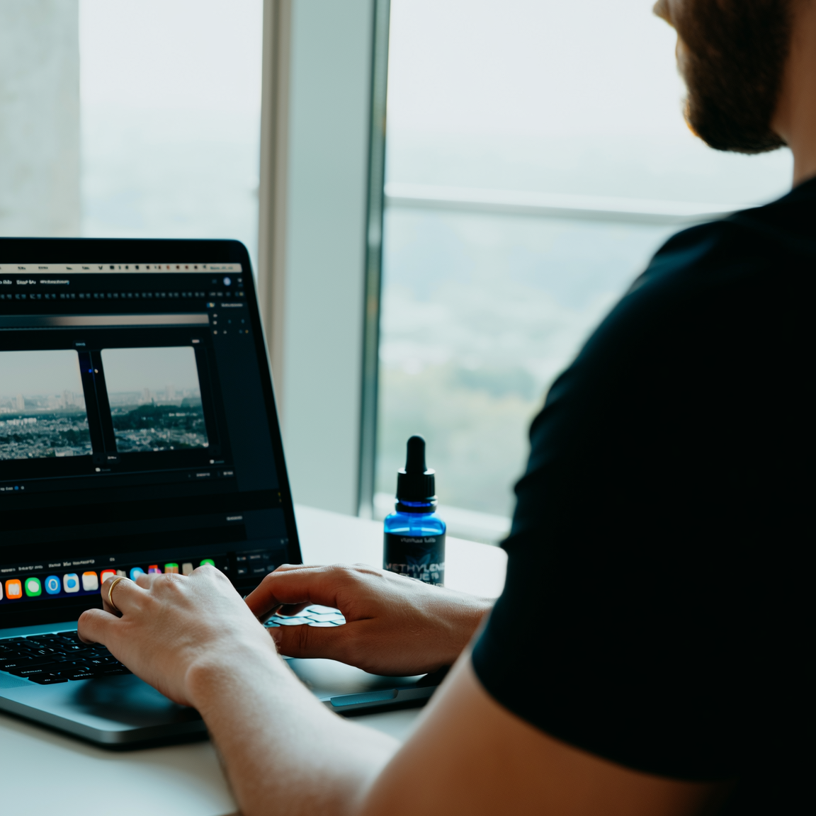 Person using a laptop at a desk with a bottle of oil on a windowsill.